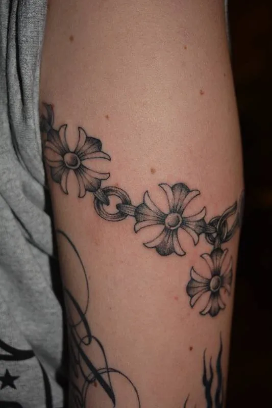 Black and grey fine-line tattoo of a daisy-chain bracelet linked by twine wrapping around the bicep, by Scripttoria at Ohana Tattoo, Waikiki, Hawaii.