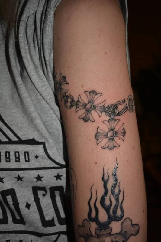 Black and grey fine-line tattoo with a daisy-chain bracelet on the upper arm and a bone-and-flame piece below it, by Scripttoria at Ohana Tattoo, Waikiki, Hawaii.