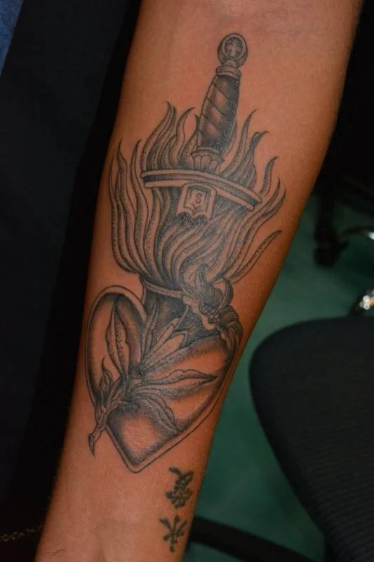 Black and grey Traditional sacred heart tattoo with a dagger driven through and flames rising, on the forearm, by Scripttoria at Ohana Tattoo, Waikiki, Hawaii.