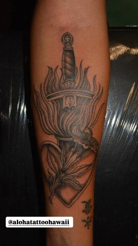Black and grey Traditional tattoo of a dagger driven down into a column of flame above a heart-shaped shovel and lily, on the calf, by Scripttoria at Ohana Tattoo, Waikiki, Hawaii.