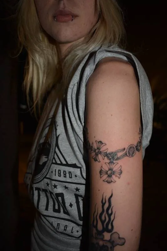 Black and grey fine-line tattoo of the daisy-chain bracelet pendant, closer crop on the outer bicep, by Scripttoria at Ohana Tattoo, Waikiki, Hawaii.