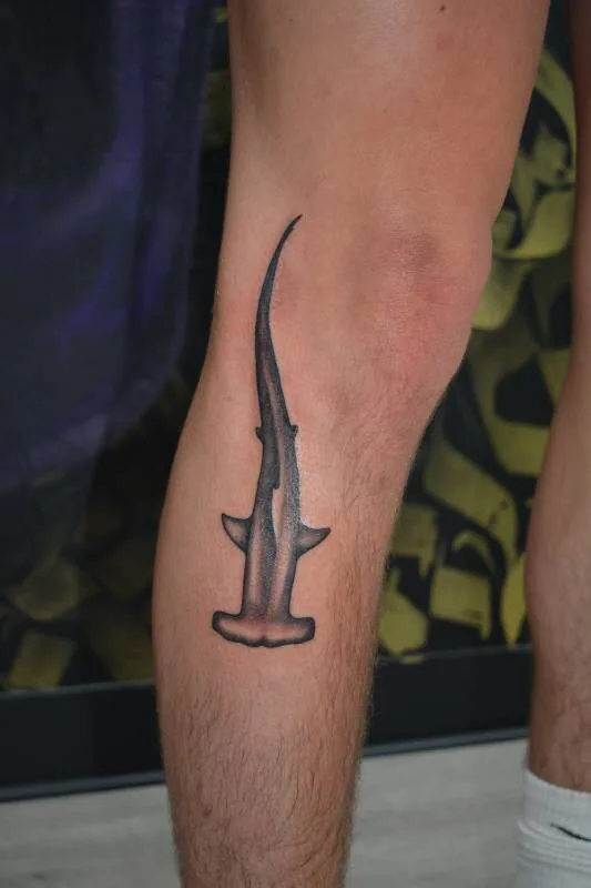 Black and grey stylized hammerhead shark silhouette tattooed vertically on the calf, by Scripttoria at Ohana Tattoo, Waikiki, Hawaii.
